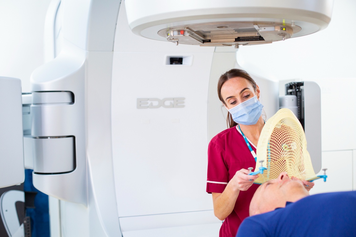 Patient lying on radiotherapy machine bed. A therapeutic radiographer is placing a mask over his head to keep him in position for radiotherapy to his head or neck