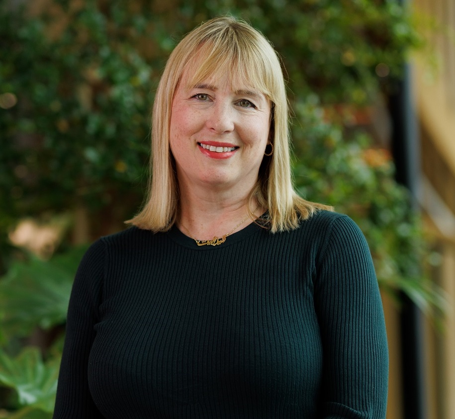 Liz is standing in a bright airy building foyer in front of a wall with live foliage growing on it. She is smiling and has shoulder length hair with a fringe.