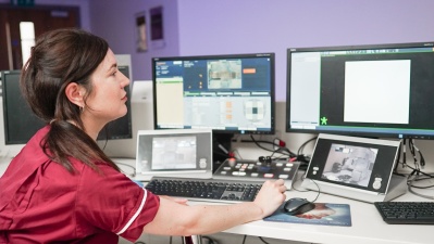 Therapeutic radiographer sitting at a control desk with screens in front of her so she can see the patient and the information about their treatment