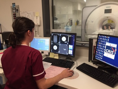 Radiographer in burgundy scrubs observing patient on scanner next door through a glass screen. She is also looking at MRI images from the scan