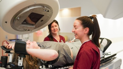 Two therapeutic radiographers in burgundy scrubs help a patient get into position on a radiotherapy machine treatment bed