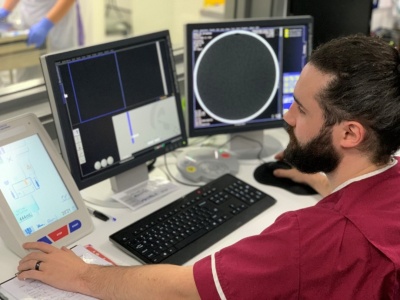 Radiographer in burgundy scrubs sitting at desk with control panel and displays from CT scanner