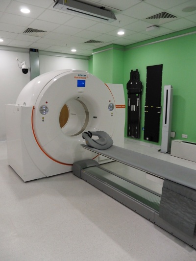 The PET-CT scanner has a flat bed that people lie on while being scanned. The scanner is a large doughnut-shaped ring that rotates around the bed to take images 