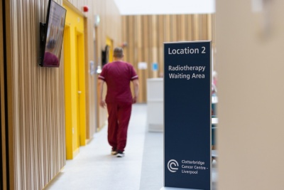 Therapeutic radiographer in burgundy scrubs walking into waiting area with light wooden panels and a sign that says Radiotherapy waiting area